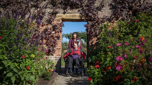 Two people, one in a wheelchair, coming through a garden gate looking at the flowers all around
